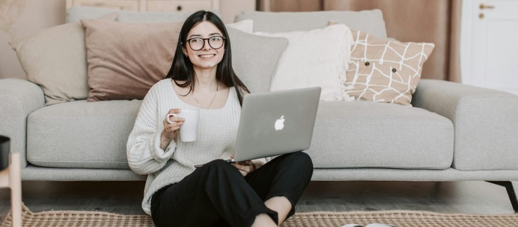 Happy woman enjoying coffee while using a laptop in a cozy living room setting.